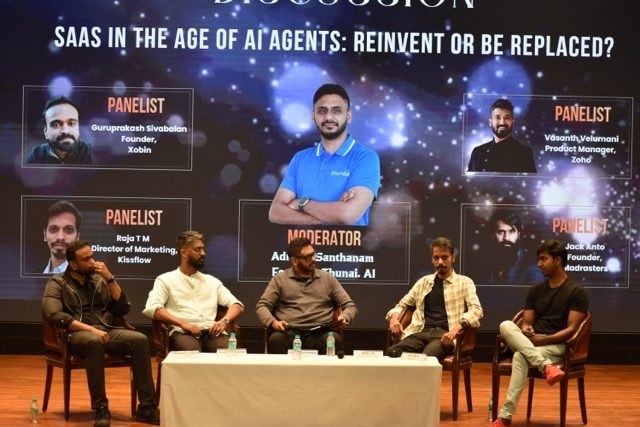 The five-person panel seated on stage beneath the screen listing the moderator and panelists